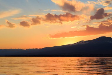 Deniz ve dağların üzerinde güzel bulutlu gün batımı. Turuncu güneşin ışınlarında parlak bir gökyüzü. Doğal