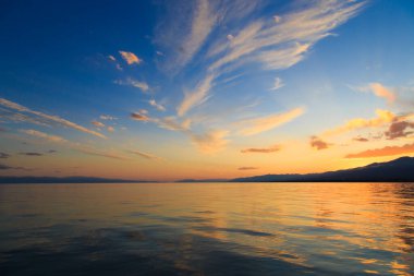 Deniz ve dağların üzerinde güzel bulutlu gün batımı. Turuncu güneşin ışınlarında parlak bir gökyüzü. Doğal