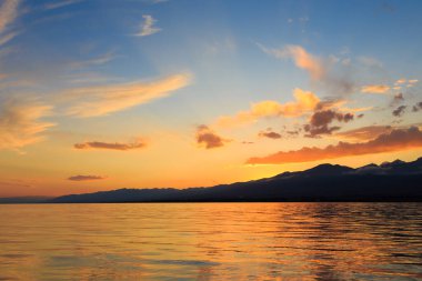 Deniz ve dağların üzerinde güzel bulutlu gün batımı. Turuncu güneşin ışınlarında parlak bir gökyüzü. Doğal