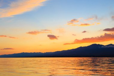 Deniz ve dağların üzerinde güzel bulutlu gün batımı. Turuncu güneşin ışınlarında parlak bir gökyüzü. Doğal