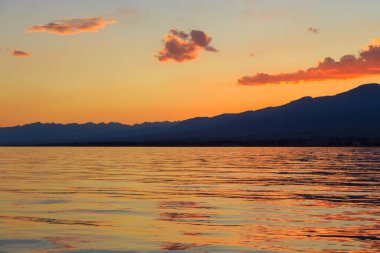 Deniz ve dağların üzerinde güzel bulutlu gün batımı. Turuncu güneşin ışınlarında parlak bir gökyüzü. Doğal