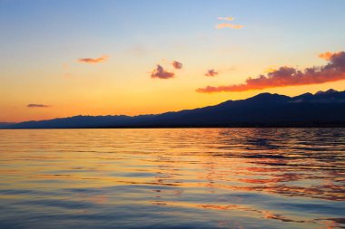 Deniz ve dağların üzerinde güzel bulutlu gün batımı. Turuncu güneşin ışınlarında parlak bir gökyüzü. Doğal
