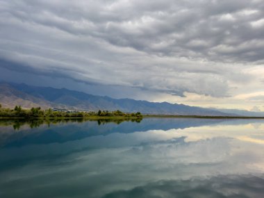 Issyk-Kul gölünün sakin suları dramatik bulutları ve dağları yansıtır. Kırgızistan 'daki doğal bir vahada sessizlik ve sükunet..