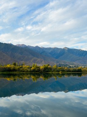 Bir dağ gölünün sakin suyu mavi gökyüzünü ve beyaz bulutları yansıtır. Yeşil kıyı şeridi gölü çevreleyerek uyum ve sükunet hissi yaratıyor.