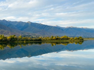Bir dağ gölünün sakin suyu mavi gökyüzünü ve beyaz bulutları yansıtır. Yeşil kıyı şeridi gölü çevreleyerek uyum ve sükunet hissi yaratıyor.