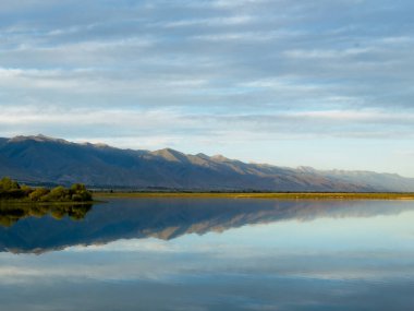 Bir dağ gölünün sakin suyu mavi gökyüzünü ve beyaz bulutları yansıtır. Yeşil kıyı şeridi gölü çevreleyerek uyum ve sükunet hissi yaratıyor.