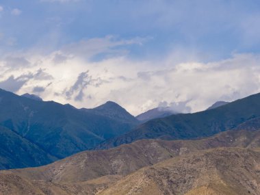 Kabarık bulutlarla dolu bir gökyüzünün altında yeşil ve kahverengiye boyanmış görkemli dağların manzarası. Engebeli arazi, yukarıdaki dingin gökyüzü ile çok güzel tezat oluşturuyor.