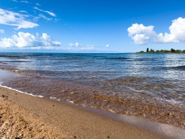 Issyk-Kul sahilinde güneşli bir sabah. Gök mavisi su, hafif bulutlar, sahil güzel kum ve çakıl taşlarıyla kaplı, hoş bir deniz esintisi.
