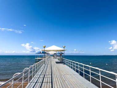 Mavi gölün üzerine uzanan beyaz gölgelikli bir iskele, kabarık bulutlarla dolu açık mavi bir gökyüzünün altına kadar uzanıyor. Sahilde mükemmel bir gün, güneş ışığında ışıl ışıl suyla.