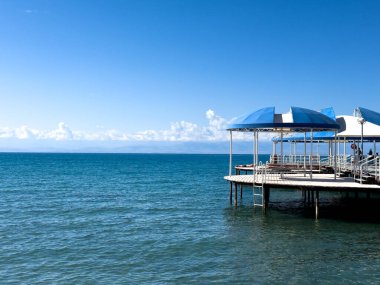 Gölün mavi-yeşil suları üzerinde mavi gölgelikleri olan küçük beyaz bir iskele uzanır. Parlak güneşli bir günde deniz bulutsuz gökyüzünü yansıtır.