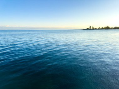 Issyk-Kul Gölü 'nün arka planında bulutlu bir gökyüzü ve dağlar var. Su sakin, gökyüzünü yansıtıyor. Sabahın erken saatlerinde doğanın dinginliği ve huzuru