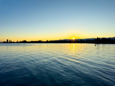 Issyk-Kul Gölü 'nün arka planında bulutlu bir gökyüzü ve dağlar var. Su sakin, gökyüzünü yansıtıyor. Sabahın erken saatlerinde doğanın dinginliği ve huzuru