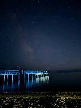 Yıldızlı bir gökyüzünün altında gölün karanlığına açılan uzun bir iskele. Sessiz gece, parlayan gökyüzü, ve sönük uzak ışık huzurlu ve mistik bir atmosfer yaratır.