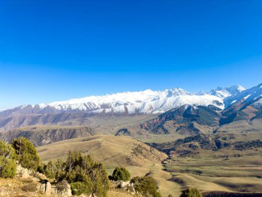 Kırgızistan 'daki karla kaplı dağ sıralarının panoramik manzarası açık mavi gökyüzünün altında, yeşil vadiler ve dağ eteklerindeki sonbahar ormanları.