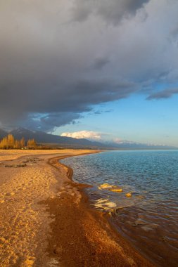 Kırgızistan, Issyk-Kul Gölü kıyısında gün batımı, yumuşak kumlu plajlar ve dağlara karşı bulutlu bir gökyüzü.