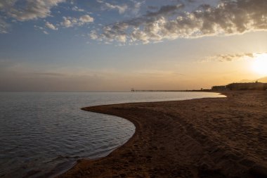 Kırgızistan 'daki Issyk-Kul Gölü' nün sakin sularında gün batımı, kumlu bir sahil ve sessiz bir ufuk.