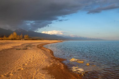 Kırgızistan, Issyk-Kul Gölü kıyısında gün batımı, yumuşak kumlu plajlar ve dağlara karşı bulutlu bir gökyüzü.