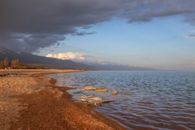 Kırgızistan, Issyk-Kul Gölü kıyısında gün batımı, yumuşak kumlu plajlar ve dağlara karşı bulutlu bir gökyüzü.