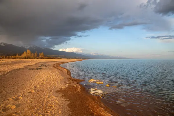 Kırgızistan, Issyk-Kul Gölü kıyısında gün batımı, yumuşak kumlu plajlar ve dağlara karşı bulutlu bir gökyüzü.