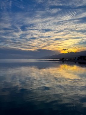 Sessiz dağ gölü, akşam güneşi, Kırgızistan. Gölde durgun su ve dağ yansıması var. Issyk-Kul 'un üzerinde gün batımı sakin sulardaki yansımalarla