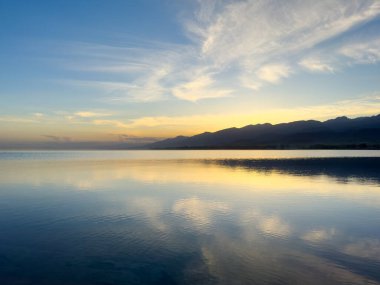Sessiz dağ gölü, akşam güneşi, Kırgızistan. Gölde durgun su ve dağ yansıması var. Issyk-Kul 'un üzerinde gün batımı sakin sulardaki yansımalarla