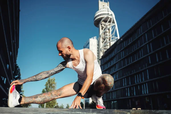 Young muscular tattooed man doing exercises, stretching early in the morning, bald bearded guy in sportswear stretching legs before jogging