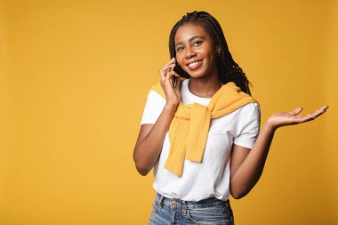 Portrait view of good looking young adult woman calling friend, talking on cell phone with calm expression. Indoor studio shot isolated on yellow background