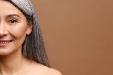 50 'li yaşlarda, neşeli, orta yaşlı, gri uzun saçlı, çekici bir bayanın kameraya bakıp gülüşü. Kahverengi üzerinde çıplak omuzları olan olgun bir model.