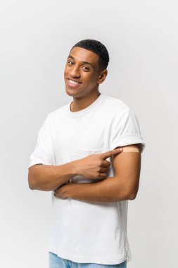 Gülümseyen genç Latin adam aşı sonrası koluna yara bandı yapıştırıyor mavi arka planda izole edilmiş, Afro-Amerikalı adam ön kolundaki dikey portreyi işaret ediyor.