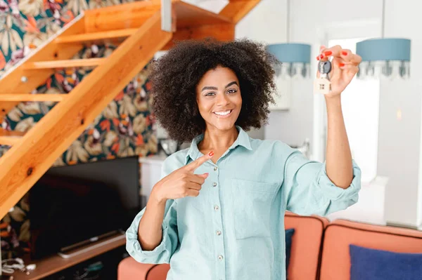 Happy with relocation woman points finger at keys with cute keychain little house, excited female student move in own dwelling, african-american relocated, moving into a new house, got a mortgage
