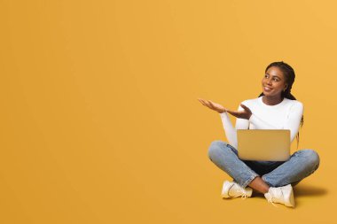 Pozitif gülümseyen serbest çalışan kadın bilgisayarla oturuyor ve boş bir yeri işaret ediyor. Tatmin olmuş çok ırklı kadın sarı arka planda izole edilmiş yeni bilgisayar uygulaması reklamı yapıyor.