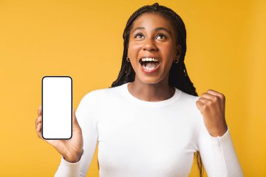 Surprised young woman using smartphone indoors for playing video game, showing him screen to the camera. Amazed happy lady rejoices, received good news, win online, raising fist up