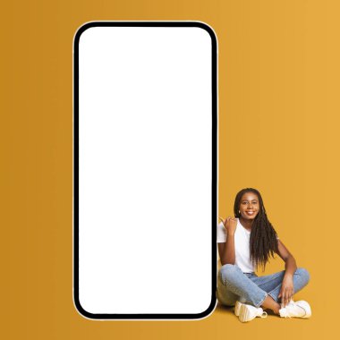 Smiling attractive girl sitting near huge smartphone with empty blank screen and showing on it with smile. Hipster woman advertising new app or website, mockup