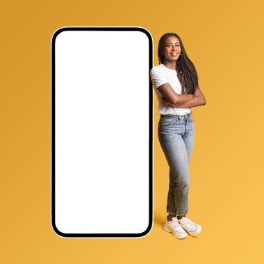 Smiling attractive girl standing near huge smartphone with empty blank screen and looking at the camera with smile. Hipster woman advertising new app or website, mockup