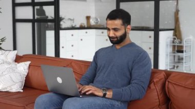 Kanepeye oturup dizüstü bilgisayar kullanan pozitif etnik adamın ön görüntüsü. Genç Hintli erkek öğrenci webinars izliyor, eğitim kursları, uzaktan öğreniyor, e-postalar yazıyor.