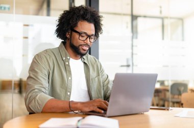 Focused curly handsome man using laptop computer in contemporary coworking space, freelance programmer in glasses coding sitting at the office desk, creativity designer or architect typing