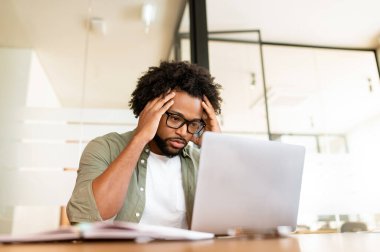 Focused on solving tasks african-american freelance guy staring at laptop screen sitting in the office, thoughtful businessman researching markets, trades stocks