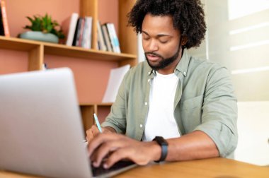 Focused and concentrated african-american male student, office worker using laptop for web searching, web surfing. Curly purposeful man in casual shirt making notes in notebook, researching