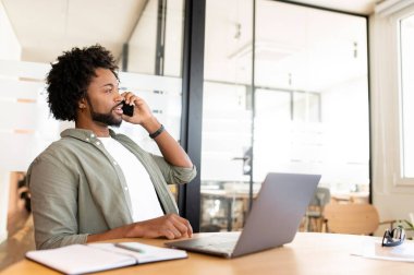 Friendly smiling african-american male employee has pleasant phone conversation sitting in modern office space, cheerful businessman in green shirt talking on smartphone