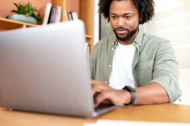 Handsome young businessman is using laptop indoors. African-American curly guy office employee is typing on the keyboard, replying to emails, male freelancer in casual wear working remotely