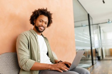 Handsome young african-american freelancer man in green casual shirt working on laptop in coworking space, enjoying remote work and free work schedule