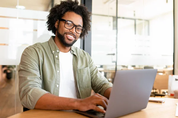 Portrait of smiling positive male office employee with computer indoors, African-American guy using laptop for remote work sitting in the modern coworking space, freelancer enjoys work on the distance