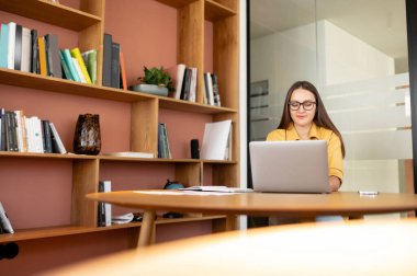 Smiling female freelancer or student sitting at the desk in office or library, studying, taking an online course, listening to the webinar, looking at laptop screen, typing