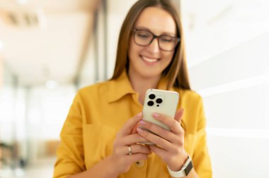 Selective focus at trendy smartphone in hands of businesswoman in eyeglasses, female office employee using phone for chatting online, messaging. Freelancer girl using phone for remotely working