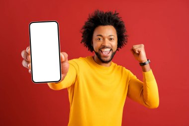 Excited hilarious African American curly young man showing smartphone with empty screen and screaming yes on red background, overjoyed guy in yellow pullover won online, celebrating victory