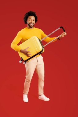 Overjoyed 30s guy holding yellow suitcase and pretend playing guitar, ready for adventure and holiday, dancing and fulling around isolated on red background, travel and vacation concept. Full length