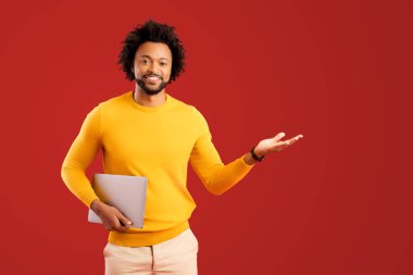 Cheerful handsome curly african-american guy hold laptop and point with palm aside at empty space isolated on plain red background, man in yellow advertising, recommending new computer app
