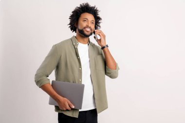 Busy handsome confident bearded african man in casual wear talking on smartphone carrying laptop isolated on white, male entrepreneur with curly afro airstyle taking a call on the mobile phone