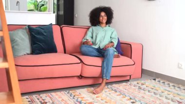 African-american woman using smartphone sitting down on comfortable couch in modern apartment, looking aside dreamly, spending lazy weekend at home