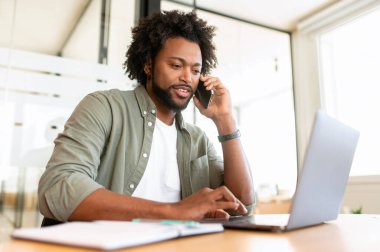 Young African-American man using laptop on the workplace and has phone conversation, male office worker chatting on the smartphone while typing on a laptop